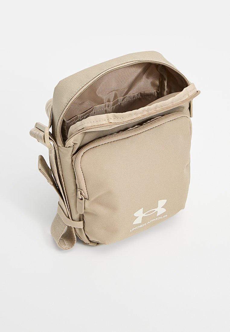 Sac messager en tissu beige avec une ouverture zippée, une sangle réglable et le logo Under Armour. Texture extérieure lisse, design compact.