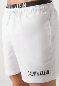 Biele plavky s elastickým čiernym pásom, na ktorom je v tučných písmenách logo značky "CALVIN KLEIN". Hladká, ľahká tkanina.