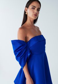 Mėlyna satino suknelė be petnešėlių, plačiomis, klostuotomis rankovėmis ir struktūruotu liemeniu. Lygi tekstūra ir plati silueto linija, papildyta papuošalais, kurie yra paprasti.