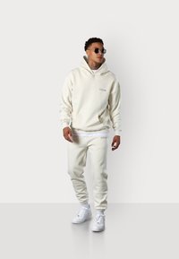 Cremefarbener Hoodie mit Fronttasche und Logo, kombiniert mit passenden Jogginghosen. Weiße Sneakers vervollständigen das Outfit. Glattes Material, lässiger Schnitt.
