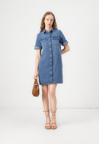 Kjole i denim med korte ermer, knappestolpe foran, to brystlommer og rett snitt, komplementert med en brun veske og sandaler med spenner.