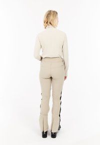 Vrouw in een beige ribbelige coltrui en beige broek met zwarte zijstrepen, staand met haar rug naar de camera op een witte achtergrond.