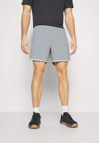 Vuxen som bär ljusgrå sportshorts, svart tröja, svarta sneakers med vita strumpor, stående mot en enkel vit bakgrund.