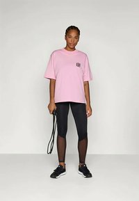 Moteris rožinės spalvos didelio dydžio marškinėliuose ir juodose leginsuose laiko juodą šokinėjimo virvę, stovėdama prieš balto fono.