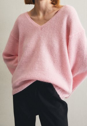 Personne portant un pull oversize doux rose pâle à col en V, assorti à un pantalon noir ample, debout devant un fond uni.