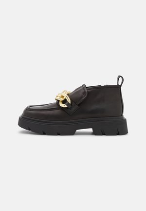 Musta nahkainen loafer-mallinen kenkä paksulla pohjalla, jossa kultainen ketjidetalji ja takana vetolenkki, näkyvissä sivulta valkoisella taustalla.