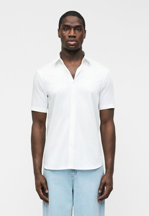 Junger Mann trägt ein schlichtes weißes kurzärmeliges Hemd mit Knöpfen und hellblaue Jeans, steht vor einem weißen Hintergrund.