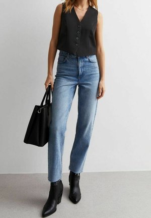 Femme portant un gilet noir sans manches boutonné, un jean bleu clair taille haute, des bottines noires, tenant un sac à main noir devant un mur blanc.