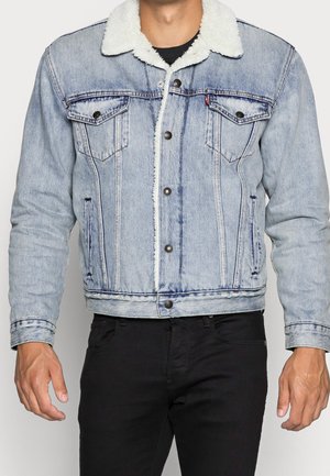 Mann trägt hellblaue Jeansjacke mit weißem Lammfellkragen und schwarze Hose, steht mit entspannten Armen an den Seiten.