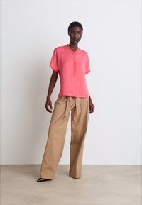 Kurze, rosa Bluse mit kurzen Ärmeln und V-Ausschnitt sowie dezenten Falten, kombiniert mit hochgeschnittenen, weiten beigen Hosen. Elegante schwarze Schuhe.