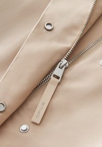 Giacca beige con zip caratterizzata da inserti metallici, texture a costine e un'etichetta in tessuto con la scritta "OPUS". Dettaglio prominente con bottoni a pressione.