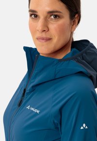 Veste imperméable bleue avec capuche, dotée d'un zip intégral, de poignets élastiques et d'accents de logo sur la poitrine et la manche. Texture lisse.