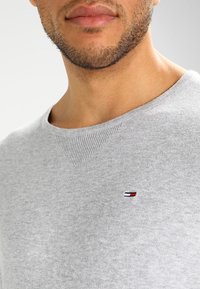 Gros plan sur la partie inférieure du visage et le cou d'un homme portant un pull ras du cou gris clair avec un petit logo rectangulaire rouge, blanc et bleu sur la poitrine.