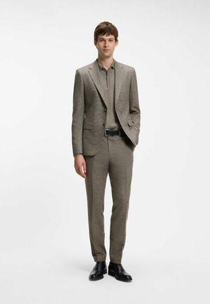 Jonge man die een bruin gestructureerd pak draagt met bijpassend poloshirt, zwarte riem en zwarte nette schoenen, staand tegen een effen witte achtergrond.