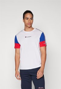 Camiseta blanca con mangas cortas azules y rojas. Presenta un logo de Champion en el pecho y un diseño a rayas a lo largo de las mangas.