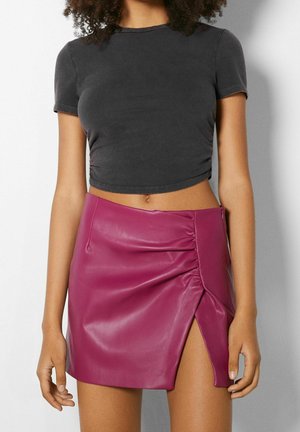 Femme portant un t-shirt court noir et une mini-jupe en similicuir magenta avec une fente latérale et des détails froncés.