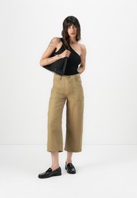 Top nero a spalla unica, pantaloni cropped khaki con tasche frontali e mocassini neri. La modella tiene una borsa nera su uno sfondo bianco.
