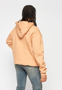 Sudadera con capucha de color durazno claro, de corte holgado y mangas largas, con un diseño liso y puños acanalados, combinada con jeans azules.