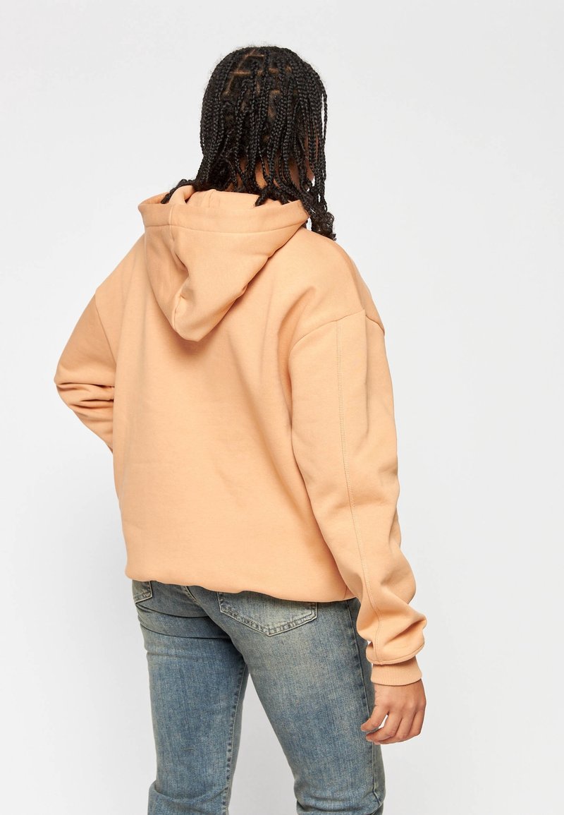 Sudadera con capucha de color durazno claro, de corte holgado y mangas largas, con un diseño liso y puños acanalados, combinada con jeans azules.