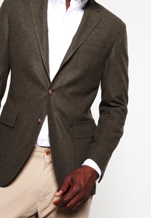 Dunkelgrüner Blazer mit einem strukturierten Karomuster, zwei vorderen Taschen und einem Ein-Knopf-Verschluss, getragen über einem weißen Hemd.
