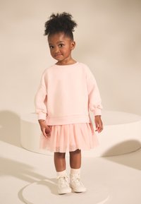 Világosrózsaszín pulóver pufó ujjakkal, hozzá réteges tüllszoknya. Fehér tornacipővel egészül ki a szett, egy kör alakú emelvényen állva.