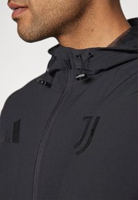 Giacca nera con cappuccio testurizzato, cordino e chiusura con zip. Loghi neri distintivi sul petto e sulla spalla sinistra.
