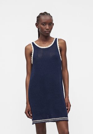 Vrouw met gevlochten haar die een mouwloze marineblauwe gehaakte jurk draagt met witte geschulpte rand op een effen achtergrond.