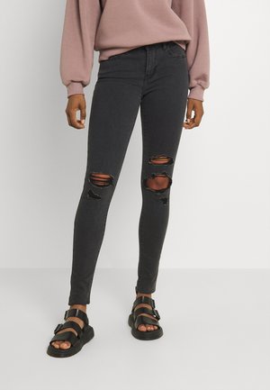 Person, die schwarze, zerrissene Skinny-Jeans, ein mauvefarbenes Langarmshirt und schwarze Sandalen mit Schnalle trägt und vor einem weißen Hintergrund steht.