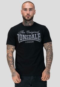 Mees, kellel on täislapi tätoveeringud ja kaelatätoveering, kannab musta "Lonsdale London" t-särki, seistes ühetaolise heleda halli taustal.