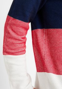 Gestreepte gebreide trui met marineblauwe, rode en witte panelen en een gestructureerd patroon. De mouw heeft een strakke pasvorm en geribbelde manchetten.