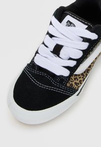 Sneaker noir et blanc avec des lacets blancs, panneau latéral en imprimé léopard et semelle en caoutchouc blanche sur un fond clair.