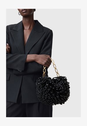 Schwarze Handtasche mit einem strukturierten Design und Schlaufen, ergänzt durch einen goldenen Kettenhenkel. Getragen mit einem schwarzen Blazer, ohne sichtbare Muster.