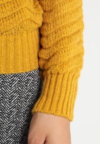 Rukáv žlutého žebrovaného svetru s texturovaným vzorem pletení. Kombinován s černobílou látkou v kostičkovaném vzoru, která zobrazuje kontrastní detaily.