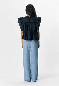 Schwarzes Oberteil mit Puffärmeln und gerafftem Taillendetail; kombiniert mit hellblauen weit geschnittenen Jeans. Glatte Textur, schlichtes Design und keine sichtbaren Beschläge.