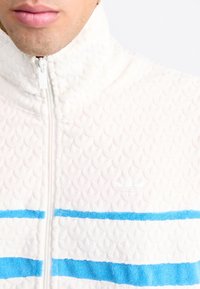 Bílá zipovací bunda s prošívanou strukturou, s modrými horizontálními pruhy a malým bílým logem na přední straně.