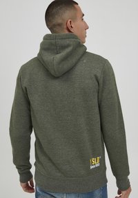 Donkergroen hoodie met een capuchon, ribgebreide manchetten en onderrand. Bevat gele en witte geborduurde tekst aan de onderrand. Zachte, gestructureerde stof.