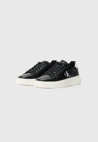 Sneaker in pelle nera con suola di gomma bianca, dotate di punta arrotondata, lacci frontali e un logo sul lato. Dettagli testurizzati sulla suola.