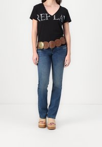 Camiseta negra de cuello en V con letras grises, jeans azules con ligero desgarro y un cinturón de cuero marrón con detalles redondeados y herrajes dorados.