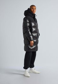 Cappotto puffo nero con finitura lucida, cappuccio oversize e polsini a costine. Indossato sopra pantaloni della tuta neri e scarpe da ginnastica bianche.