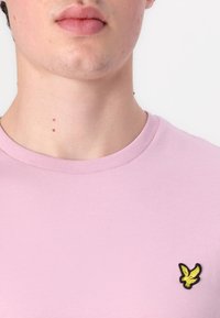 Világos rózsaszínű póló, kerek nyakkivágással, a bal mellrészén egy kis sárga hímzett saslogóval. Simító, pamut anyag.