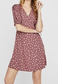 Frau, die ein mauve-farbenes Wickelkleid mit Punkten und Puffärmeln trägt, posiert mit einem Arm verschränkt und der anderen Hand nahe ihrem Gesicht.