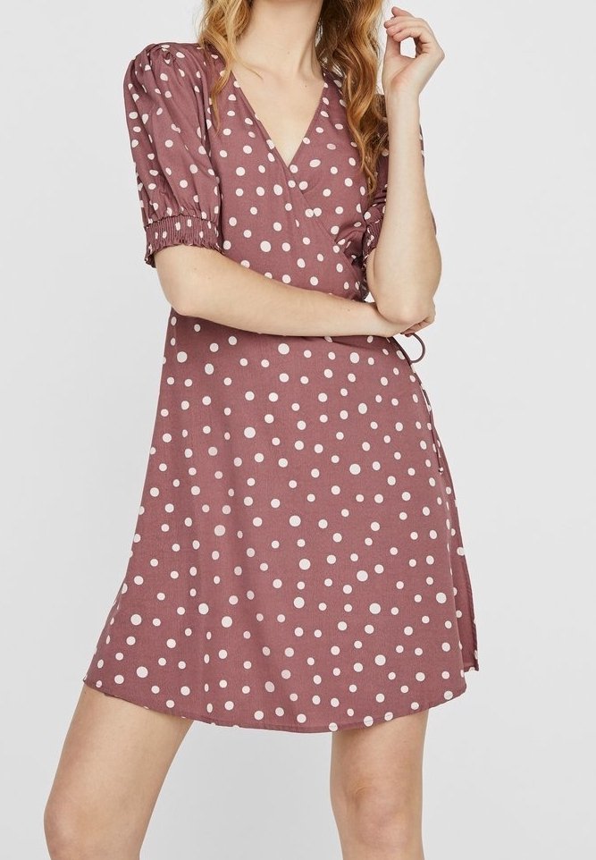 Frau, die ein mauve-farbenes Wickelkleid mit Punkten und Puffärmeln trägt, posiert mit einem Arm verschränkt und der anderen Hand nahe ihrem Gesicht.