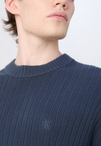 Maglione a costine in navy con collo a girocollo e un discreto logo ricamato sul petto. Tessuto texturizzato, design aderente, accenti minimi.