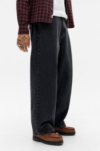 Oseba v ohlapnih črnih trapericah, rjavih mokasinah, rdeči karirasti srajci in beli majici, stoji ob enotni ozadju.