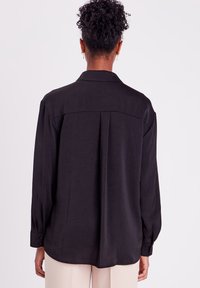 Chemise noire à manches longues en tissu doux. Présente un col classique, des poignets boutonnés, un pli au dos et une coupe décontractée avec un ourlet arrondi.
