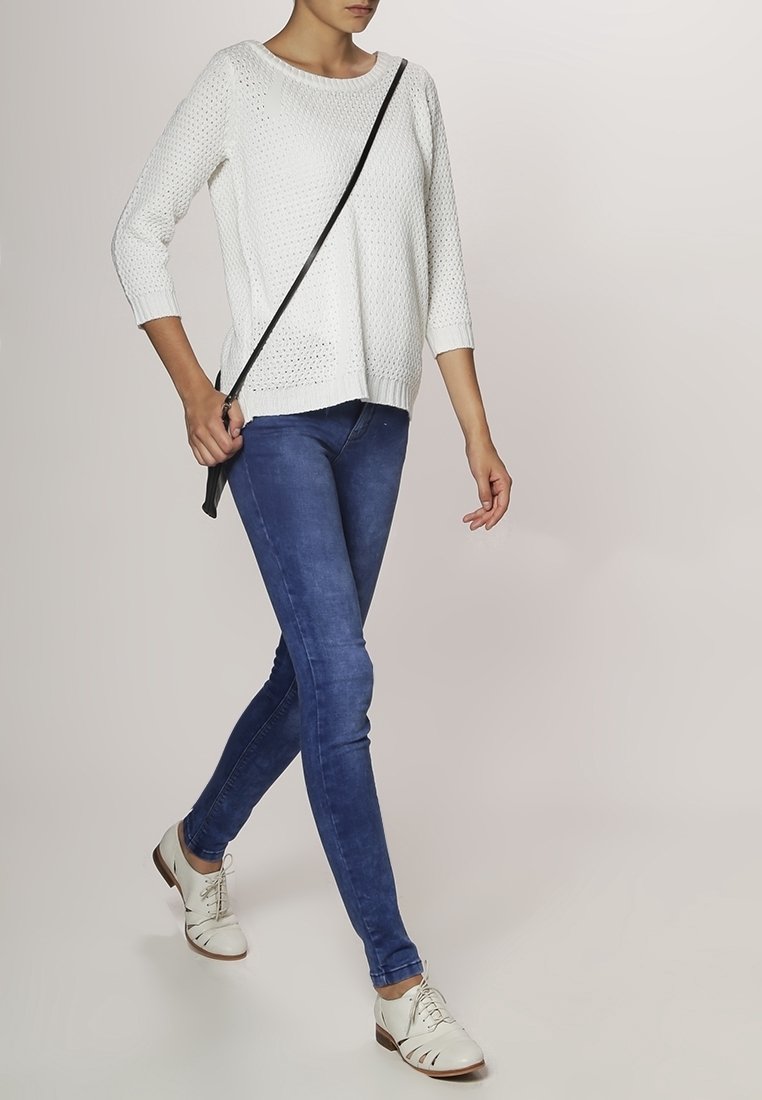 Witte gebreide trui met een losse pasvorm, blauwe skinny jeans en witte veterschoenen. Accessoires zijn een zwarte schoudertas.