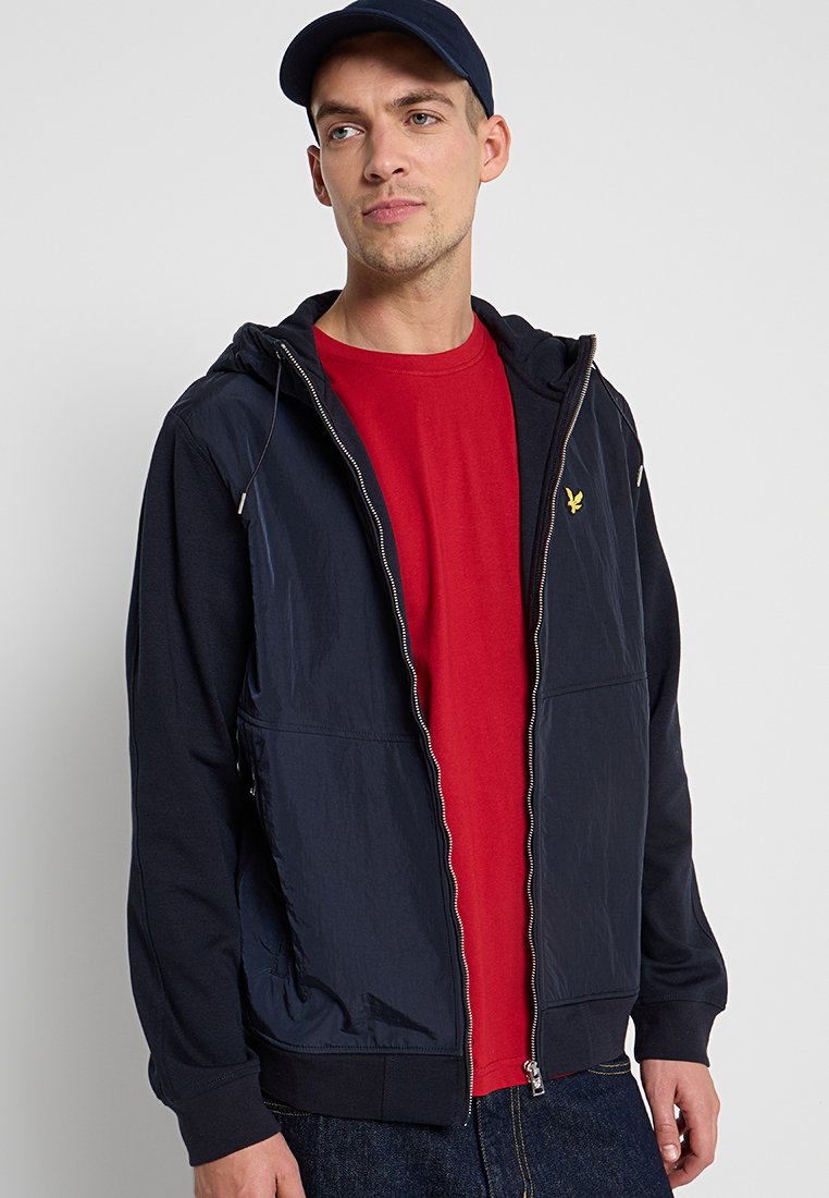 Veste zippée marine avec capuche, fabriquée à partir de matériaux mixtes. Présente un intérieur rouge, des poches latérales et un petit logo sur la poitrine.