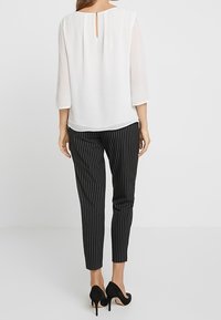 Weiße Bluse mit Dreiviertelärmeln, Rückenschlitzdetails, kombiniert mit schwarz gestreiften Hosen und schwarzen High Heels.