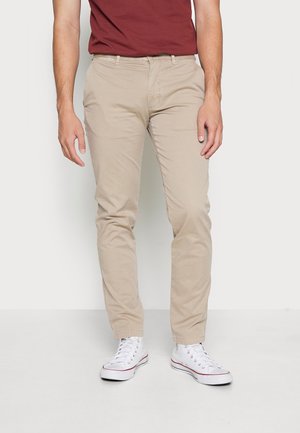 Mand iført beige slim-fit bukser, hvide høje sneakers og en mørkerød t-shirt, stående mod en ensfarvet lys baggrund.