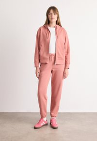 Lichtroze sweatshirt met capuchon, ritsluiting en voorzakken, gecombineerd met bijpassende trainingsbroek. Onder het sweatshirt een witte T-shirt en roze sneakers.